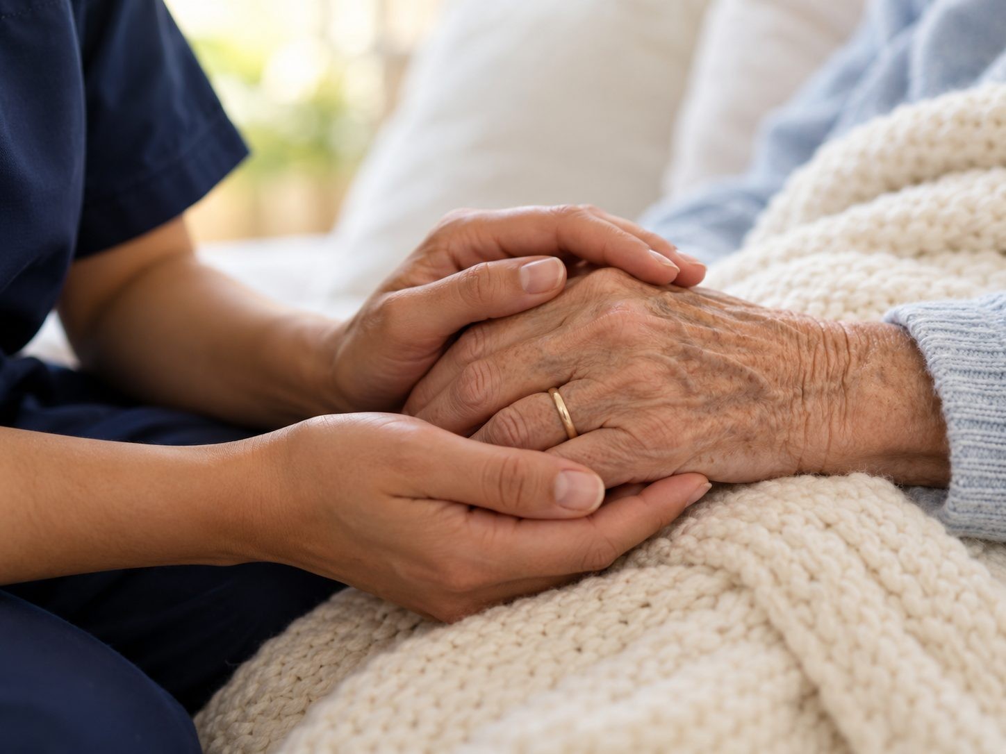A nurse's hands gently holding an elderly person's hand on a knitted blanket.