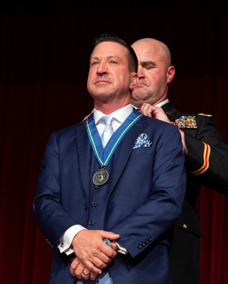Rich Johnson, founder of Cairo Hospice, stands in a navy suit as a soldier in dress uniform places the Ohio Veterans Hall of Fame medal around his neck.